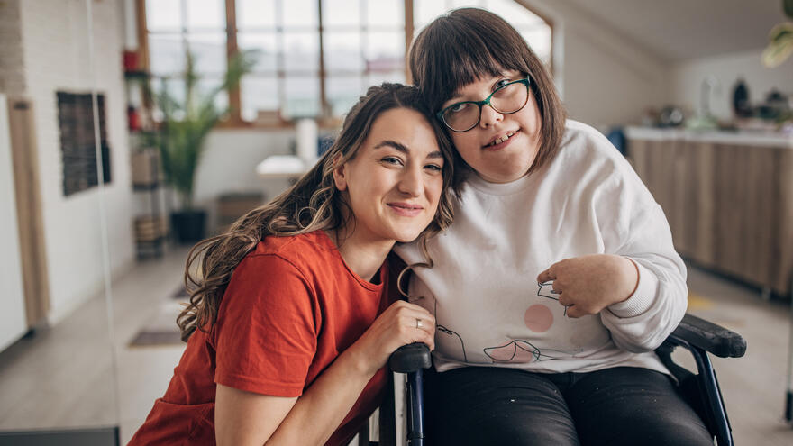 A women in a wheelchair smiling together with her friend who is knelling next to her and leaning in. 
