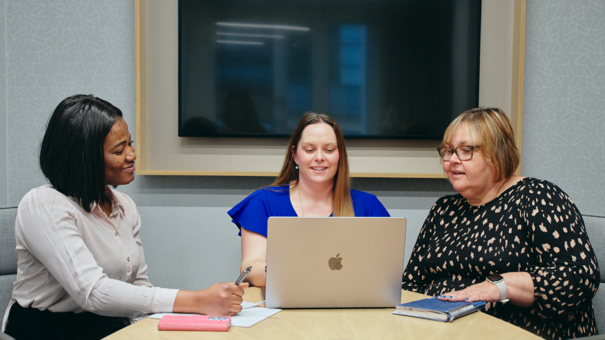 Three women sat together at a desk, looking at an open laptop computer than is positioned in front of them.