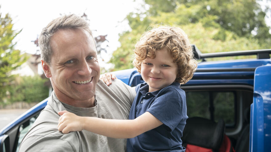 A father holding his young son in his arm, after just getting him out of the car, smiling and looking happy. 