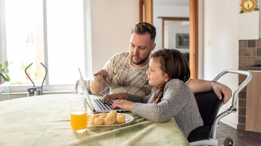 A father sat at a table pointing at something on the laptop to his daughter who is sat beside him in a wheelchair. 