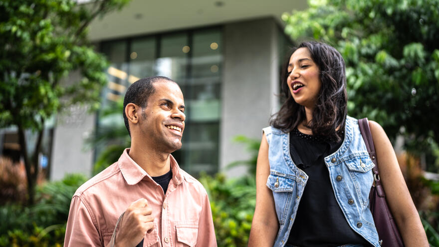 A visually impaired man holding a white cane stood together with a young woman laughing and talking to one another.