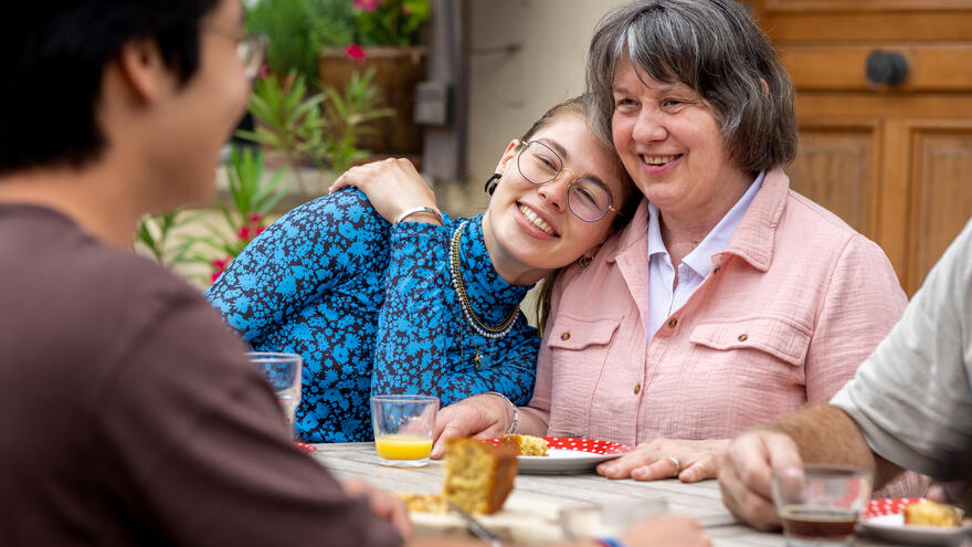 A young woman sat resting her head affectionately on the should of her mum who is sat beside her. They are relaxed and both smiling. 