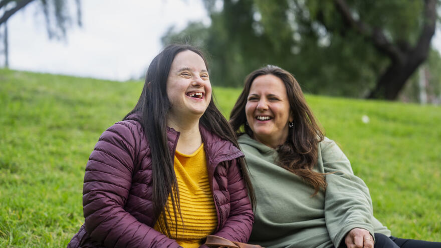 Two women sat beside one another on the grass in a park who are laughing together. 