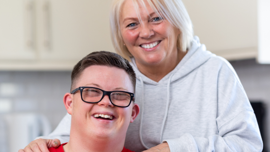 A mum proudly posing with her son with both of her hands resting on top of his shoulders who is sat in front of her. 