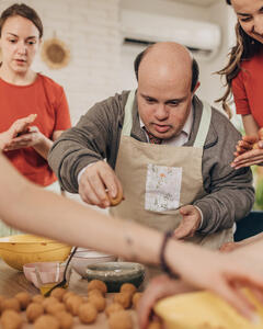 A man learning to cook in a kitchen with help and direction from 2 woman. 